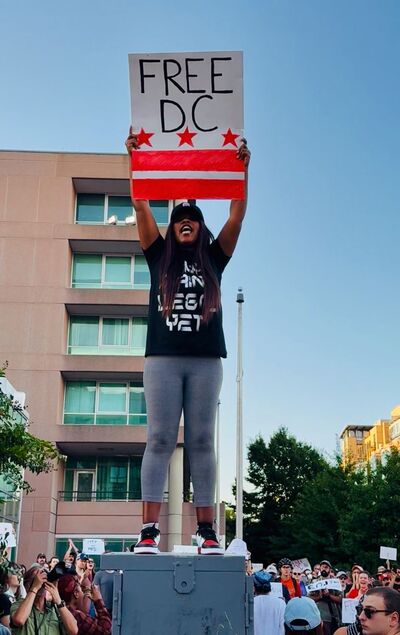 A woman stands on a box at a protest, holding a sign that says "FREE DC" with the DC flag. She is wearing a black t-shirt, gray leggings, and red shoes. A crowd of protestors is visible below her.